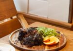 A wooden table set for a meal with a large, rustic wooden platter of barbecued meat topped with seasoning, alongside sliced oranges and a green vegetable side. In the foreground, there are place settings with plates, napkins, and cutlery. - Hausion