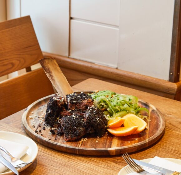 A wooden table set for a meal with a large, rustic wooden platter of barbecued meat topped with seasoning, alongside sliced oranges and a green vegetable side. In the foreground, there are place settings with plates, napkins, and cutlery. - Hausion