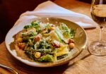 A plate of Caesar salad, topped with croutons and grated cheese, is set on a wooden table beside a glass of white wine. A fork, knife, and white napkin are placed to the left. The background includes part of a chair and warm, ambient lighting. - Hausion