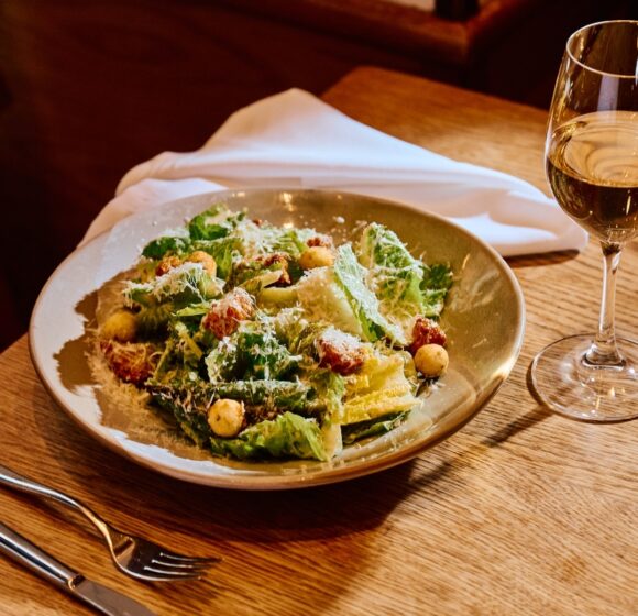 A plate of Caesar salad, topped with croutons and grated cheese, is set on a wooden table beside a glass of white wine. A fork, knife, and white napkin are placed to the left. The background includes part of a chair and warm, ambient lighting. - Hausion