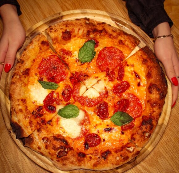 A person with red nail polish is holding a wooden tray with a freshly baked pepperoni pizza. The pizza has melted cheese, slices of pepperoni, and basil leaves, all on a slightly charred crust. The scene is set on a wooden table. - Hausion