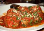 A white plate filled with meatballs covered in tomato sauce, garnished with chopped parsley and grated parmesan cheese. A spoon rests in the background on the plate. The dish is situated on a wooden surface. - Hausion