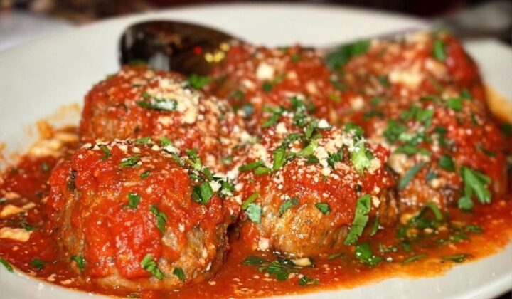 A white plate filled with meatballs covered in tomato sauce, garnished with chopped parsley and grated parmesan cheese. A spoon rests in the background on the plate. The dish is situated on a wooden surface. - Hausion