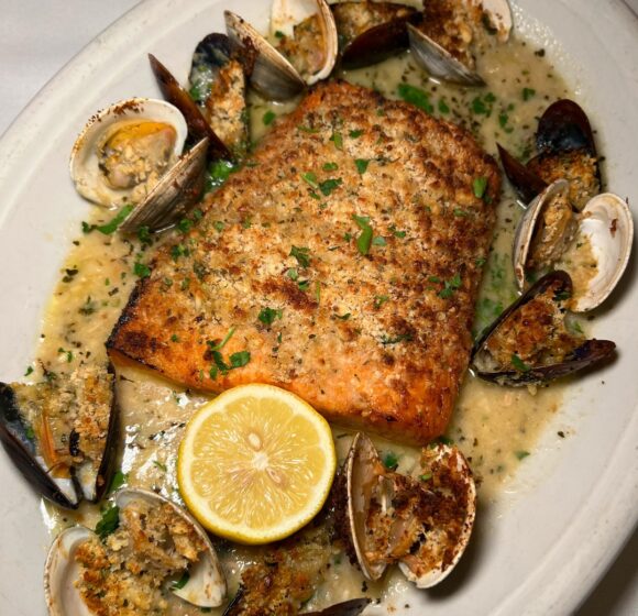 A dish featuring a roasted salmon fillet garnished with chopped herbs, served with clams and mussels in a creamy sauce. The plate is adorned with a halved lemon in the foreground. The seafood is displayed around the salmon. - Hausion