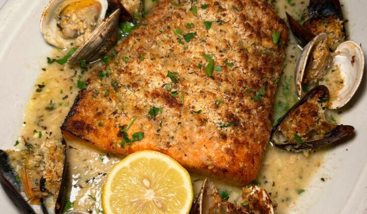 A dish featuring a roasted salmon fillet garnished with chopped herbs, served with clams and mussels in a creamy sauce. The plate is adorned with a halved lemon in the foreground. The seafood is displayed around the salmon. - Hausion