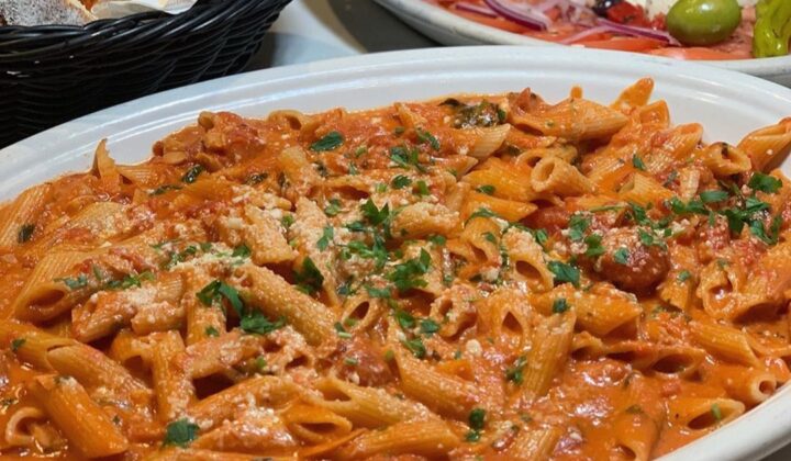 A delicious spread of Italian cuisine features a large oval dish filled with penne pasta in a rich, creamy tomato sauce, garnished with fresh herbs. In the background, there's a basket of sliced bread and a platter with sliced tomatoes, mozzarella, and olives. - Hausion