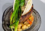 A gourmet dish featuring a piece of seared meat, topped with vibrant green leafy vegetables and a clamshell, served on a bed of chickpeas and diced vegetables. The dish is elegantly plated on a gray ceramic plate. - Hausion