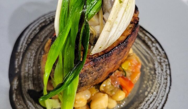 A gourmet dish featuring a piece of seared meat, topped with vibrant green leafy vegetables and a clamshell, served on a bed of chickpeas and diced vegetables. The dish is elegantly plated on a gray ceramic plate. - Hausion