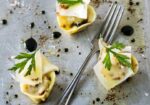 Three pieces of gourmet tortellini garnished with cheese shavings, black sauce, greens, and spices are arranged around a fork on a metal surface. Splashes of black sauce and salt crystals add visual interest to the composition. - Hausion