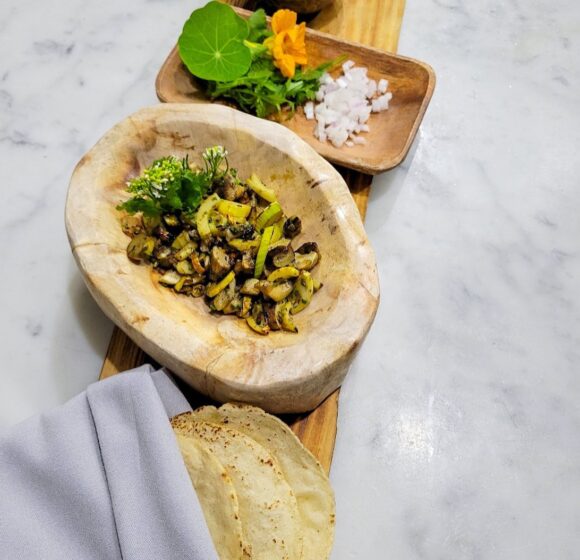 A wooden serving board holds a rustic bowl with grilled vegetables, a small plate with finely chopped onions and greens with an orange flower, a bowl of seasoning, and a neatly wrapped set of tortillas. The board rests on a light marble surface. - Hausion