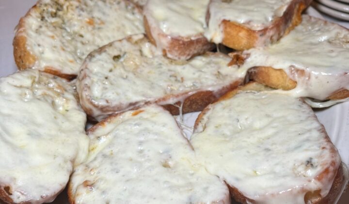 A plate of toasted bread slices generously topped with melted cheese. The bread appears crispy around the edges, and the cheese is golden and slightly bubbly. In the background, there is a glimpse of another dish and a stack of small white plates. - Hausion