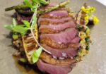 A plated gourmet dish featuring slices of seared duck breast arranged on a bed of grains, garnished with asparagus and fresh greens. The presentation is artistic, with a touch of sauce elegantly drizzled around the ingredients. - Hausion