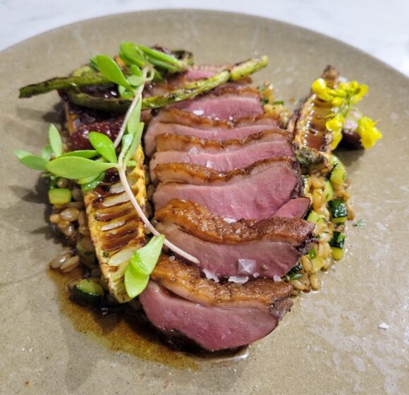 A plated gourmet dish featuring slices of seared duck breast arranged on a bed of grains, garnished with asparagus and fresh greens. The presentation is artistic, with a touch of sauce elegantly drizzled around the ingredients. - Hausion