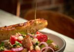 A close-up of a Mediterranean salad with tomatoes, tuna, onions, capers, and olives in a bowl on a table. A hand is drizzling olive oil over the salad, with a toasted slice of bread resting on the rim of the bowl, and a blurred background adding depth to the scene. - Hausion