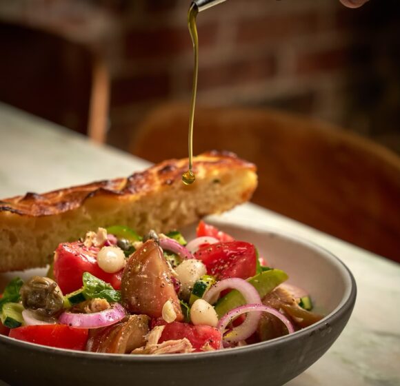 A close-up of a Mediterranean salad with tomatoes, tuna, onions, capers, and olives in a bowl on a table. A hand is drizzling olive oil over the salad, with a toasted slice of bread resting on the rim of the bowl, and a blurred background adding depth to the scene. - Hausion