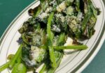 A white oval plate with charred leafy greens topped with grated cheese, placed on a green surface. The greens are slightly wilted with some crispy edges, garnished with a light dusting of cheese. The dish appears simple and fresh, emphasizing the vegetables. - Hausion