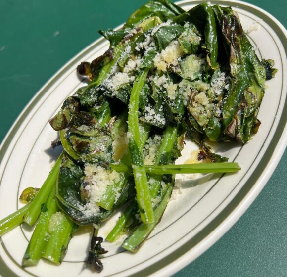 A white oval plate with charred leafy greens topped with grated cheese, placed on a green surface. The greens are slightly wilted with some crispy edges, garnished with a light dusting of cheese. The dish appears simple and fresh, emphasizing the vegetables. - Hausion