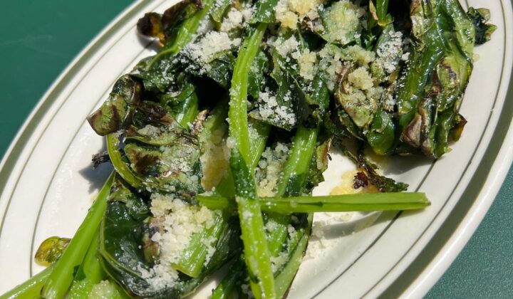 A white oval plate with charred leafy greens topped with grated cheese, placed on a green surface. The greens are slightly wilted with some crispy edges, garnished with a light dusting of cheese. The dish appears simple and fresh, emphasizing the vegetables. - Hausion