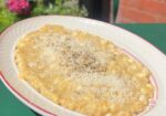 A bowl of creamy risotto topped with grated cheese and freshly ground black pepper, placed on a green table outdoors. Bright pink flowers and green foliage are blurred in the background, providing a pleasant, summery atmosphere. - Hausion