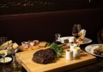 A dimly lit restaurant table set with a variety of dishes, including a large cooked steak on a wooden board garnished with herbs, a charcuterie board, assorted breads, a salad, and glasses of wine. The background features stained glass panels. - Hausion