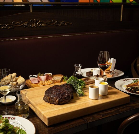 A dimly lit restaurant table set with a variety of dishes, including a large cooked steak on a wooden board garnished with herbs, a charcuterie board, assorted breads, a salad, and glasses of wine. The background features stained glass panels. - Hausion