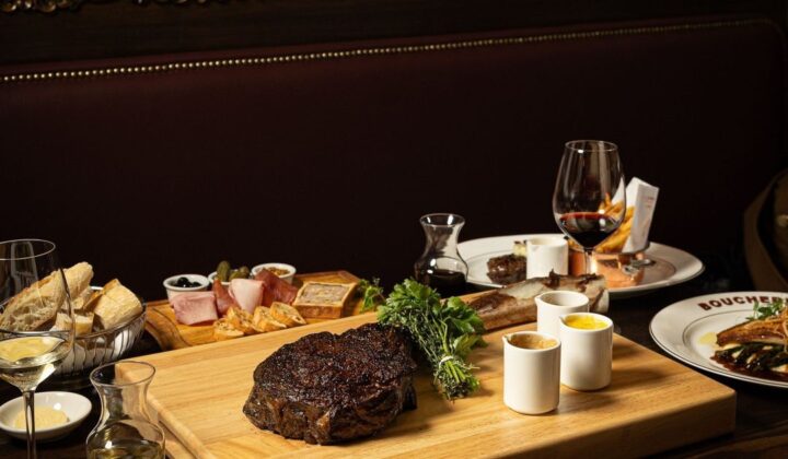 A dimly lit restaurant table set with a variety of dishes, including a large cooked steak on a wooden board garnished with herbs, a charcuterie board, assorted breads, a salad, and glasses of wine. The background features stained glass panels. - Hausion