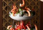 A two-tiered seafood platter featuring shrimp, lobster, clams, and mussels on a bed of ice. A hand with red nail polish and a bracelet reaches for a shrimp from the top tier. The background has a decorative fan-patterned design. - Hausion