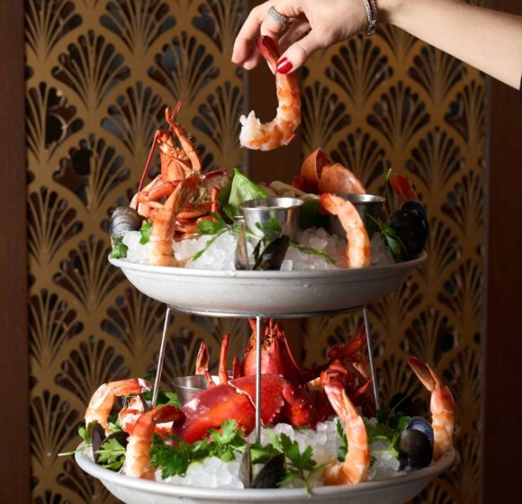 A two-tiered seafood platter featuring shrimp, lobster, clams, and mussels on a bed of ice. A hand with red nail polish and a bracelet reaches for a shrimp from the top tier. The background has a decorative fan-patterned design. - Hausion