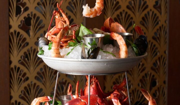 A two-tiered seafood platter featuring shrimp, lobster, clams, and mussels on a bed of ice. A hand with red nail polish and a bracelet reaches for a shrimp from the top tier. The background has a decorative fan-patterned design. - Hausion