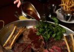 A hand pouring brown sauce over an assortment of grilled meats and bone marrow in a large pan, garnished with fresh greens. Surrounding the pan are a glass of red wine, a small jug of oil, a bowl of fries, and a silver gravy boat. - Hausion