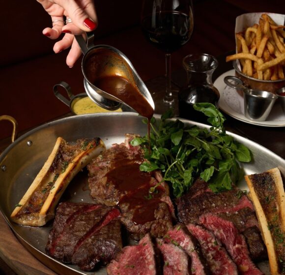 A hand pouring brown sauce over an assortment of grilled meats and bone marrow in a large pan, garnished with fresh greens. Surrounding the pan are a glass of red wine, a small jug of oil, a bowl of fries, and a silver gravy boat. - Hausion