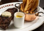 A plate of food featuring a grilled steak topped with herb butter, a serving of crispy French fries in a small copper pot, and a ramekin of yellow sauce on the side. The plate is neatly arranged on a dark wooden table. - Hausion