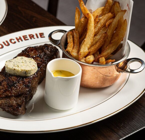 A plate of food featuring a grilled steak topped with herb butter, a serving of crispy French fries in a small copper pot, and a ramekin of yellow sauce on the side. The plate is neatly arranged on a dark wooden table. - Hausion