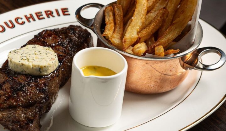 A plate of food featuring a grilled steak topped with herb butter, a serving of crispy French fries in a small copper pot, and a ramekin of yellow sauce on the side. The plate is neatly arranged on a dark wooden table. - Hausion