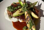 A plate of grilled steak topped with charred asparagus spears, green herb sauce, and served on a bed of creamy white sauce. The dish is garnished with additional herb sauce and sits on a neutral-colored plate with a green background. - Hausion