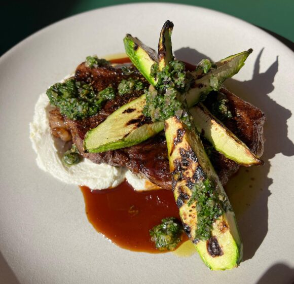 A plate of grilled steak topped with charred asparagus spears, green herb sauce, and served on a bed of creamy white sauce. The dish is garnished with additional herb sauce and sits on a neutral-colored plate with a green background. - Hausion