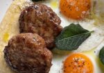 A close-up of a plate featuring two fried eggs with vibrant orange yolks, two seasoned sausage patties, fresh sage leaves, and a sprinkle of grated cheese on a creamy base. The dish is garnished with black pepper and a drizzle of olive oil. - Hausion