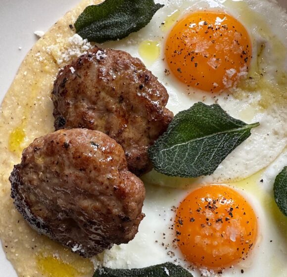 A close-up of a plate featuring two fried eggs with vibrant orange yolks, two seasoned sausage patties, fresh sage leaves, and a sprinkle of grated cheese on a creamy base. The dish is garnished with black pepper and a drizzle of olive oil. - Hausion