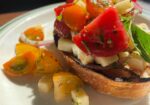 A close-up of a vibrant bruschetta dish featuring toasted bread topped with a colorful mix of chopped tomatoes, mozzarella cubes, red onions, and fresh basil. The sunny light accentuates the freshness of the ingredients. Scattered tomato pieces and herbs garnish the plate. - Hausion