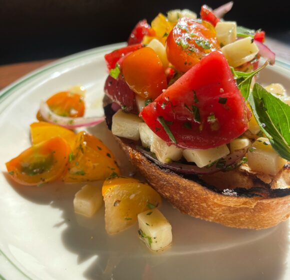 A close-up of a vibrant bruschetta dish featuring toasted bread topped with a colorful mix of chopped tomatoes, mozzarella cubes, red onions, and fresh basil. The sunny light accentuates the freshness of the ingredients. Scattered tomato pieces and herbs garnish the plate. - Hausion