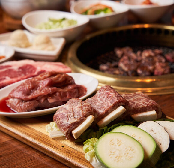 An array of raw meats and vegetables are displayed on a wooden board beside a tabletop grill. Bowls of condiments and sides surround the setup. Meat cuts include marbled beef and ribs, while vegetables include zucchini and mushrooms. The grill has cooking meat on it. - Hausion