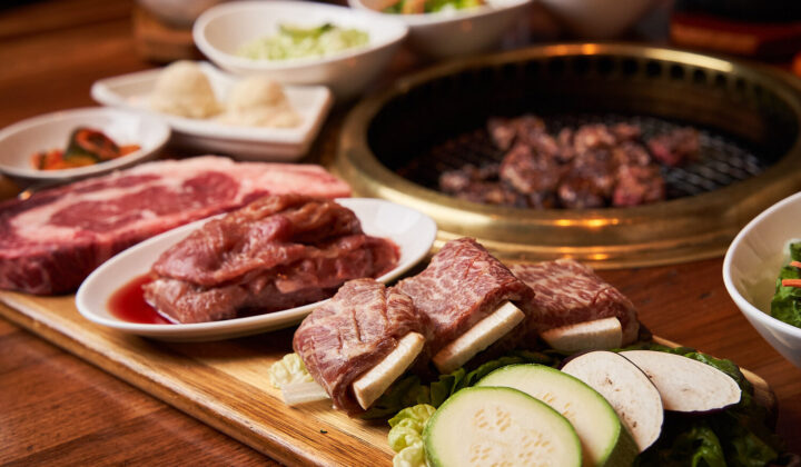 An array of raw meats and vegetables are displayed on a wooden board beside a tabletop grill. Bowls of condiments and sides surround the setup. Meat cuts include marbled beef and ribs, while vegetables include zucchini and mushrooms. The grill has cooking meat on it. - Hausion