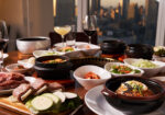 A table set with various Korean dishes including kimchi, vegetables, soups, and grilled meats, against the backdrop of a city skyline. Wine glasses and a cocktail are also present on the table. - Hausion