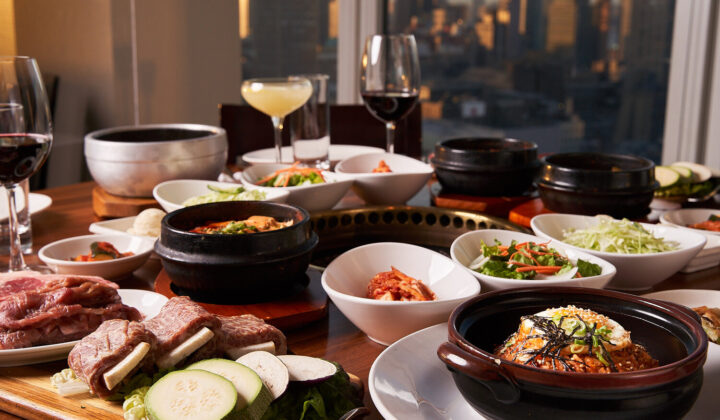 A table set with various Korean dishes including kimchi, vegetables, soups, and grilled meats, against the backdrop of a city skyline. Wine glasses and a cocktail are also present on the table. - Hausion