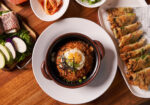 A top-down view of a meal on a wooden table. It features a bowl of Korean bibimbap with a fried egg on top, surrounded by various side dishes including kimchi, sliced vegetables, and a plate of fried dumplings. The presentation is clean and appetizing. - Hausion