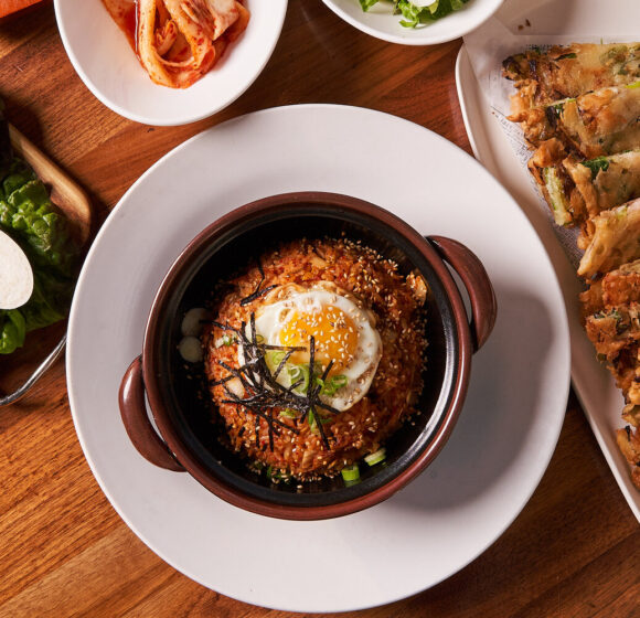 A top-down view of a meal on a wooden table. It features a bowl of Korean bibimbap with a fried egg on top, surrounded by various side dishes including kimchi, sliced vegetables, and a plate of fried dumplings. The presentation is clean and appetizing. - Hausion