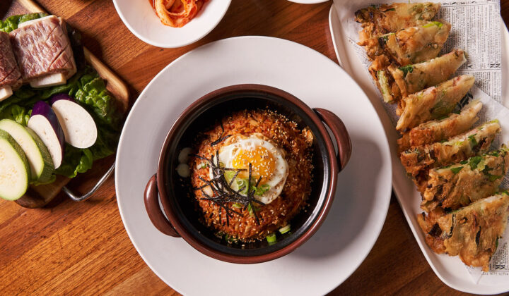 A top-down view of a meal on a wooden table. It features a bowl of Korean bibimbap with a fried egg on top, surrounded by various side dishes including kimchi, sliced vegetables, and a plate of fried dumplings. The presentation is clean and appetizing. - Hausion