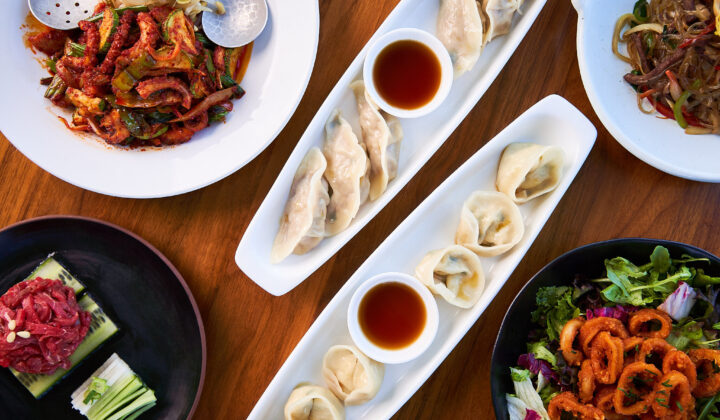 An assortment of dishes on a wooden table. The centerpiece consists of dumplings served with soy sauce in white elongated plates. Surrounding dishes include a variety of meats, vegetables, and a salad, presenting a diverse and colorful meal spread. - Hausion