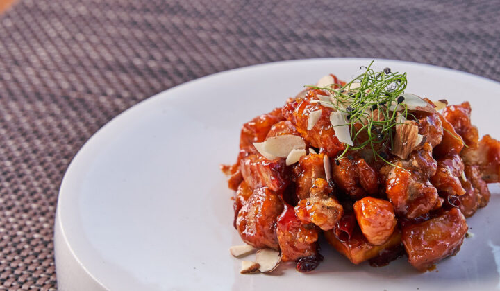 A dish of caramelized honey chicken garnished with sliced almonds and herbs is served on a white plate. The textured, brown placemat beneath the plate adds a subtle background contrast. - Hausion