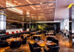 Modern restaurant interior with sleek, reflective ceiling, wooden floors, and black leather chairs around wooden tables. A long bar area with high chairs is visible to the left. Soft, warm lighting illuminates the space, creating an inviting atmosphere. - Hausion